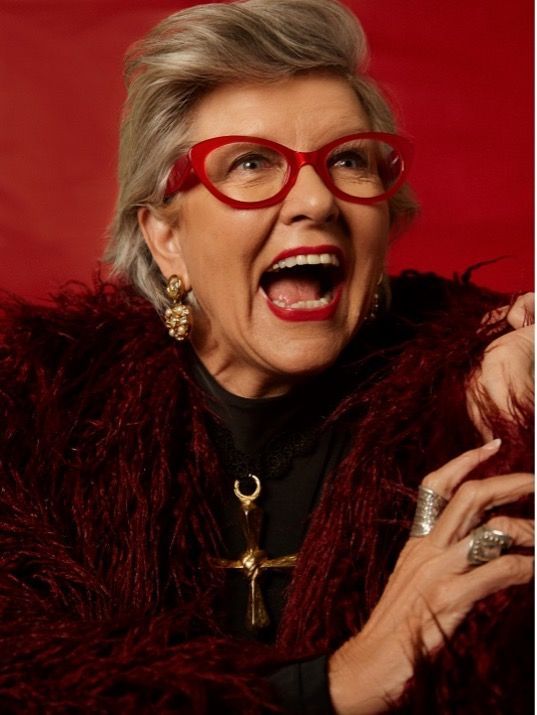 Woman with red glasses and open mouth, wearing red fur, smiling.