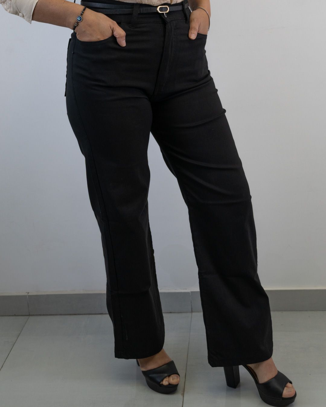Mujer con pantalones negros y tacones, con las manos en los bolsillos, de pie junto a una pared.