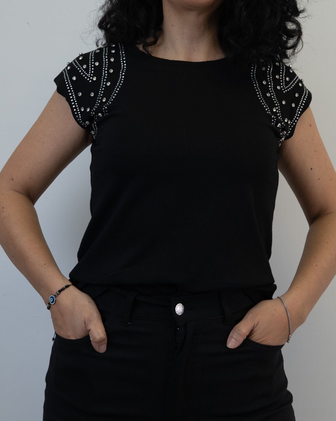 Mujer con camisa negra con hombros adornados con cuentas y manos en los bolsillos.