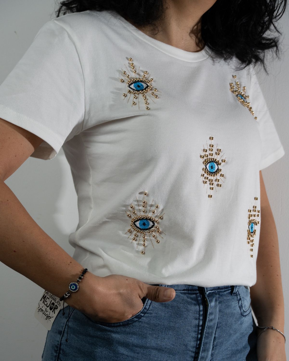 Mujer que lleva una camiseta blanca con adornos azules y dorados en forma de ojos, jeans azules y una pulsera de ojo.