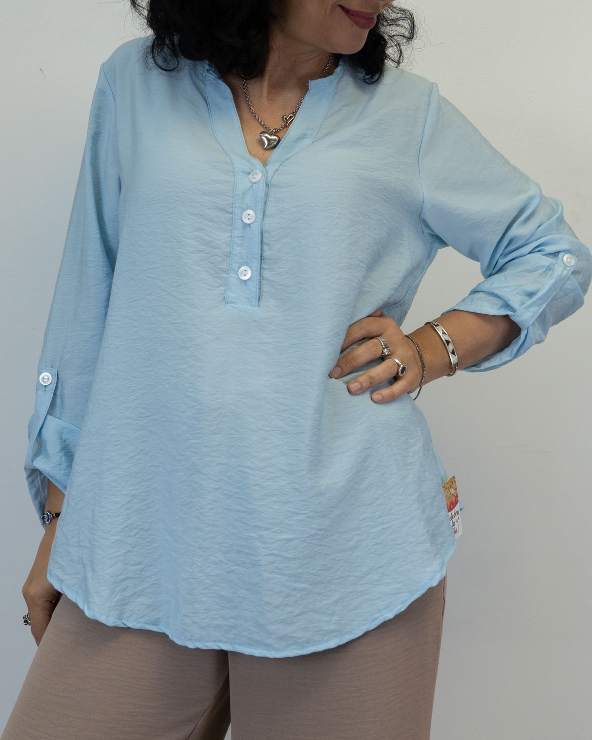 Mujer con blusa azul claro con mangas remangadas, mano en la cadera, con joyas, de pie.