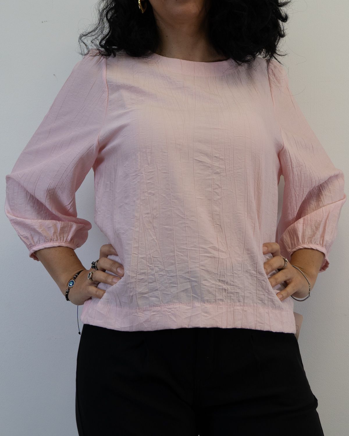 Mujer con una blusa rosa de mangas abullonadas, con las manos en las caderas y pantalones negros.