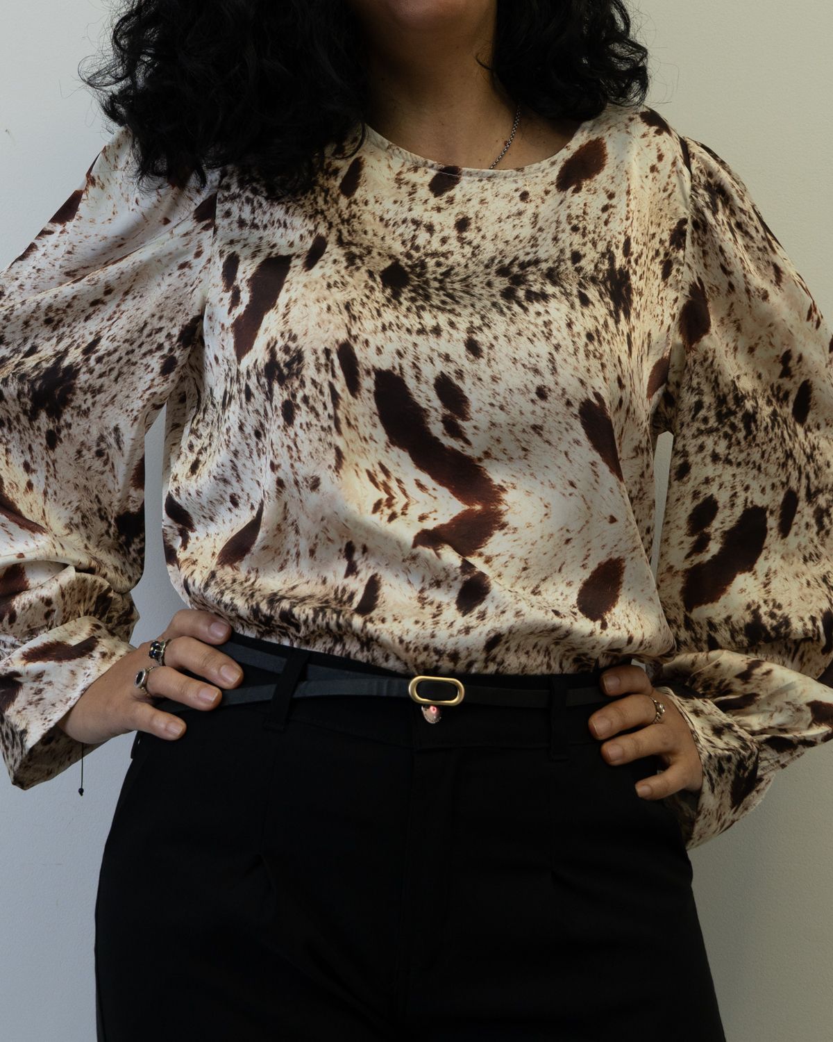 Mujer con blusa estampada con mangas abullonadas, pantalón negro y cinturón con las manos en las caderas.