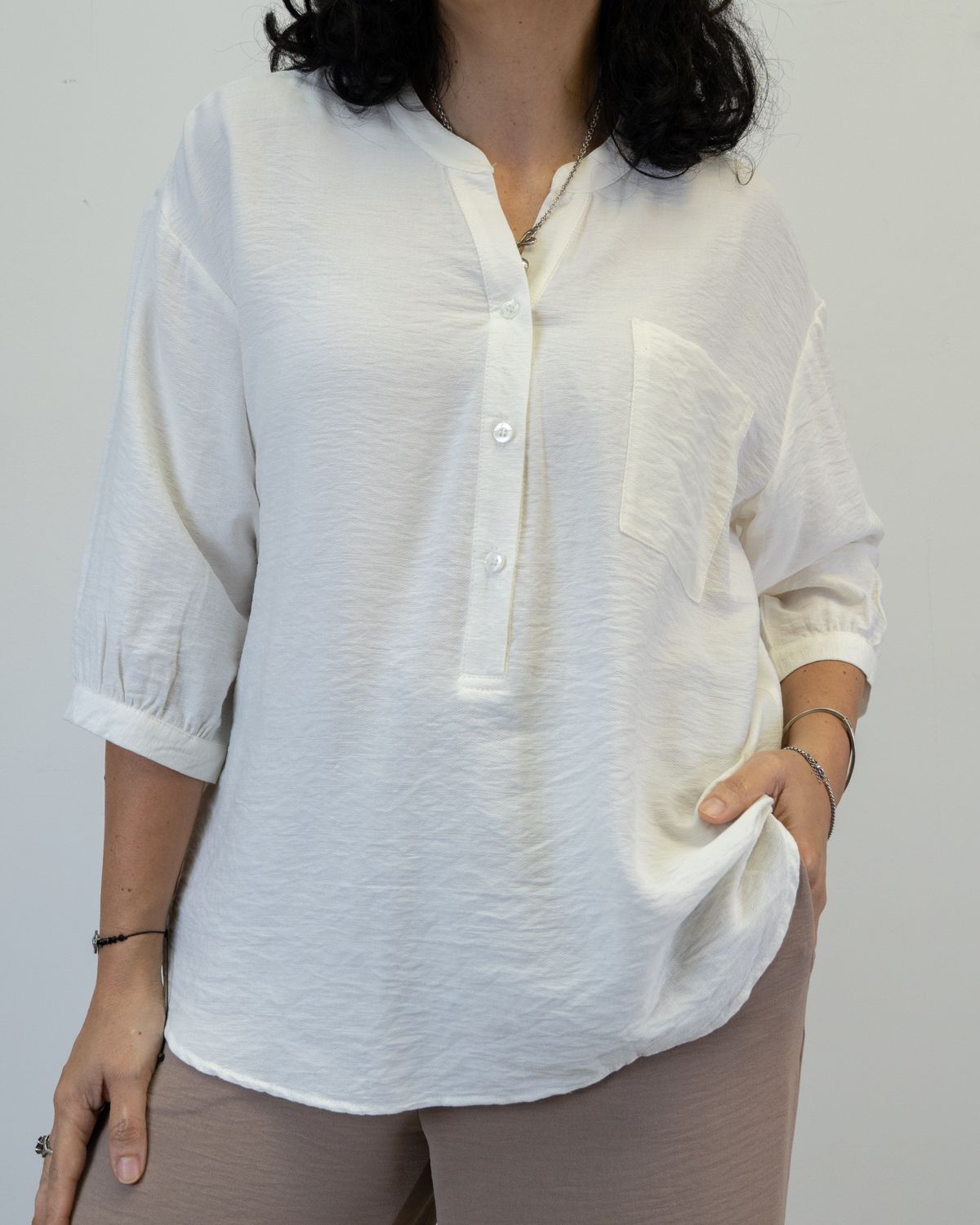 Mujer con top texturizado blanco, mangas remangadas y bolsillo, vistiendo pantalón color neutro y con la mano en el bolsillo.