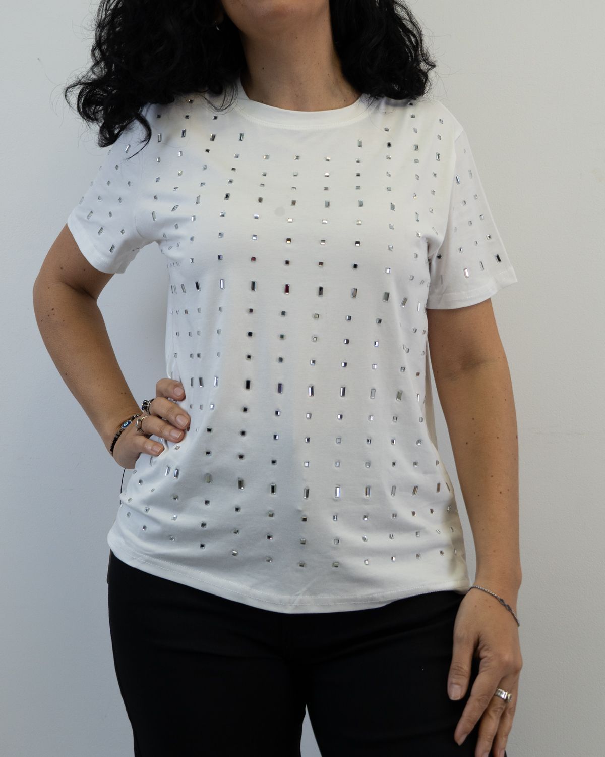 Mujer vestida con camiseta blanca con adornos plateados, pantalón negro y mano en la cadera.