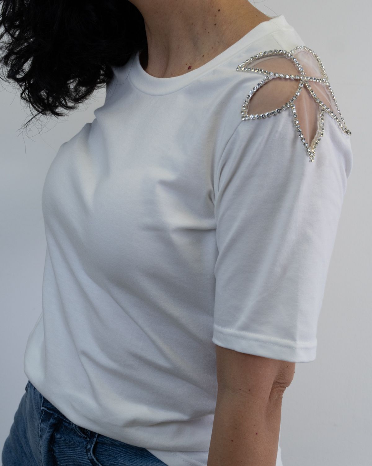 Mujer con camiseta blanca con detalle de joya en los hombros, jeans de mezclilla, de pie en el interior.