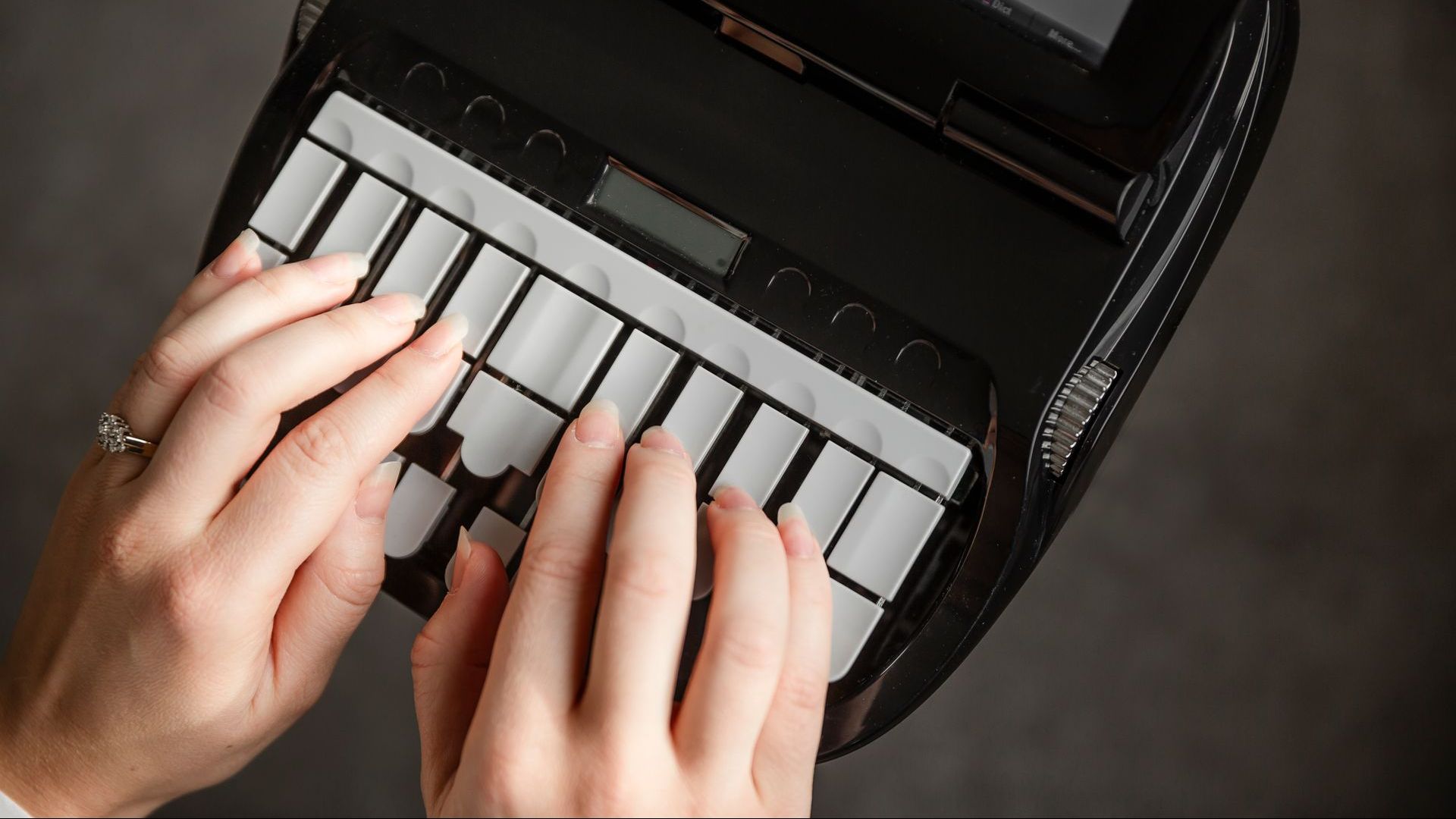 Des mains tapent sur un clavier portable noir à touches blanches. Un écran numérique est visible.