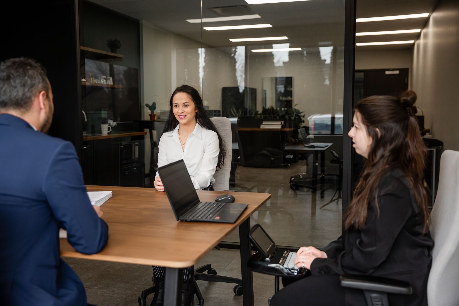 Dans un bureau moderne, trois personnes sont assises autour d'une table avec des ordinateurs portables. L'une d'elles sourit et prend la parole.
