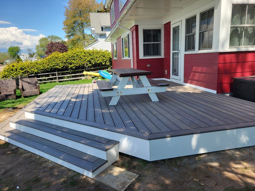 Gray deck with picnic table and steps beside a red house in a sunny yard