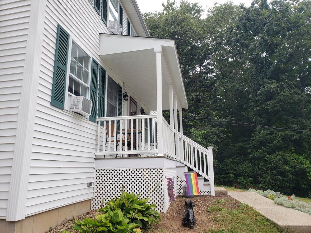 White house porch with ramp and stairs, green shutters, and a wooded yard beside a gravel path
