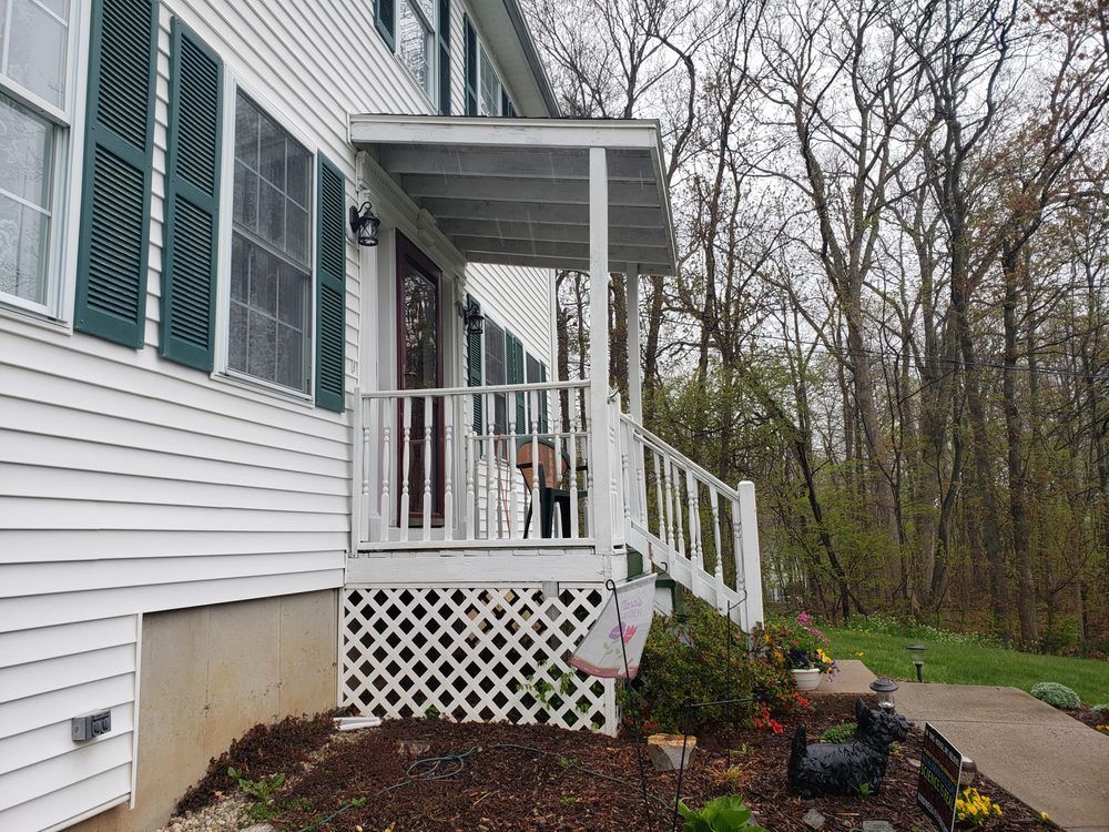 Side porch with white railings and lattice on a house, beside a wooded yard and garden bed.