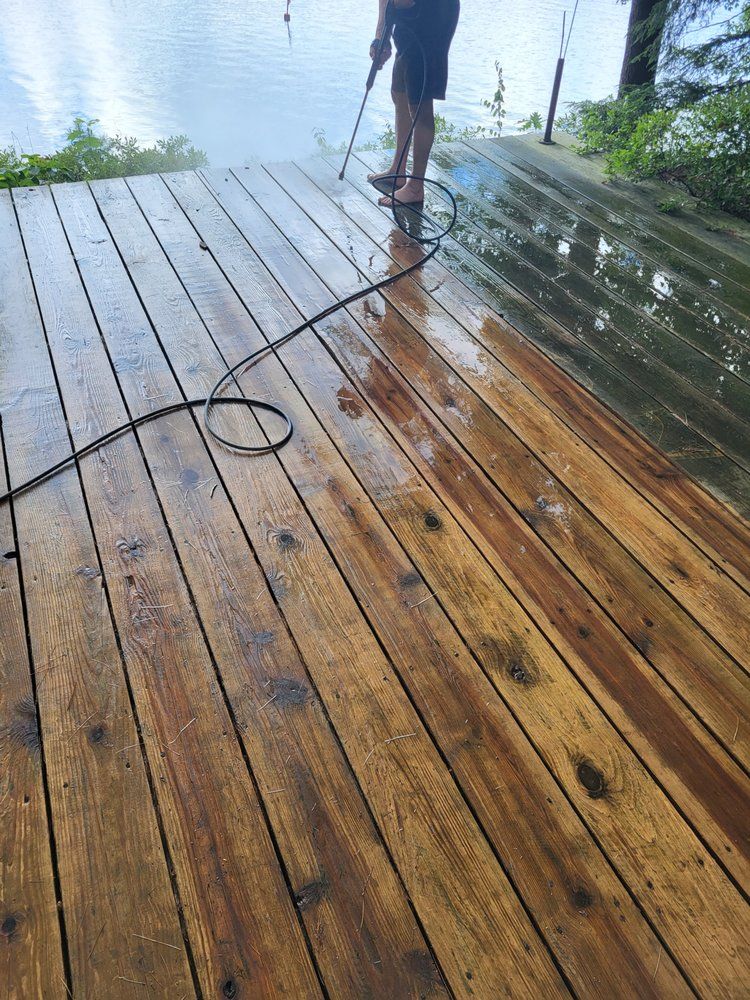 Person pressure-washing a wet wooden deck beside water, with a hose and coiled cord visible.