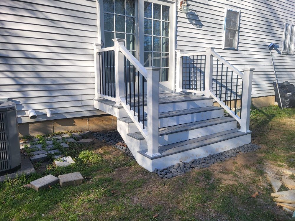 White porch steps with railings beside a gray house, leading to a small landing over gravel and grass.