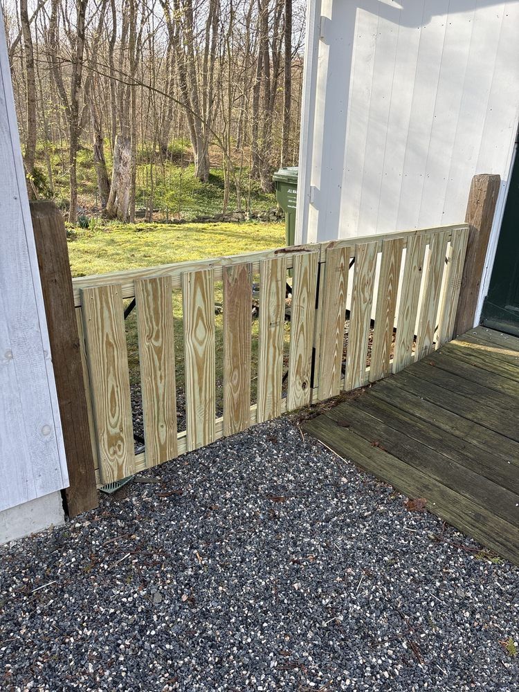 Wooden gate beside a white building, opening onto a gravel path and wooded yard.