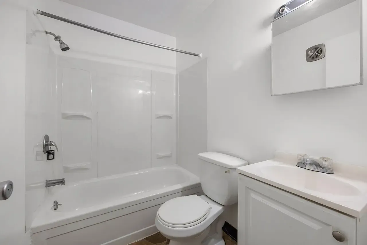 A white, brightly lit bathroom featuring a bathtub with a shower head, a standard toilet, and a vanity sink with a mirror.