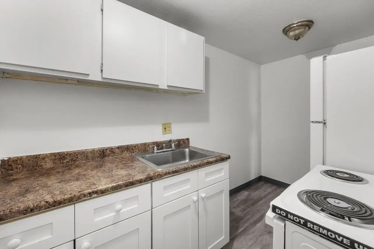 A simple kitchen featuring white cabinets, a mottled brown countertop with a stainless steel sink, and a white stove.