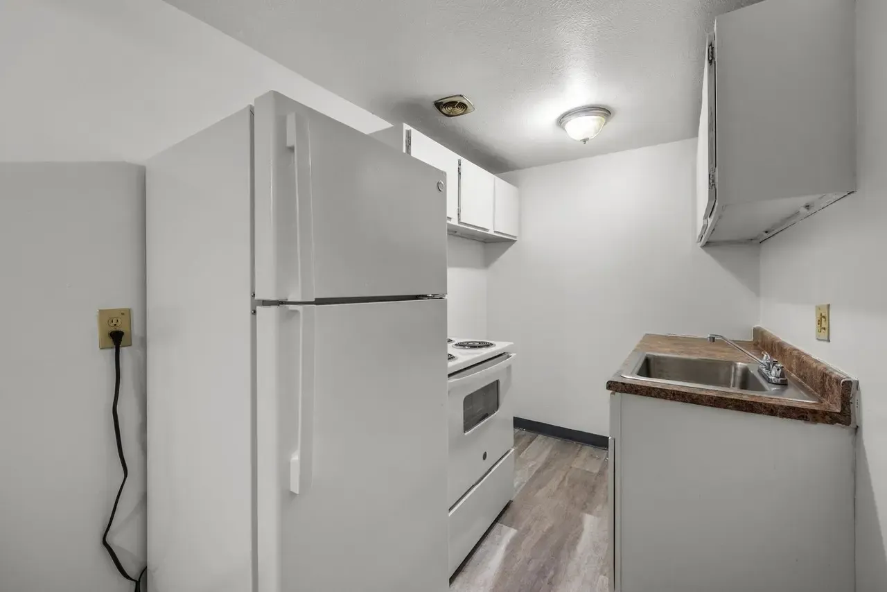 A small, white kitchen featuring a refrigerator, stove, white cabinets, and a sink with a stone-patterned countertop.