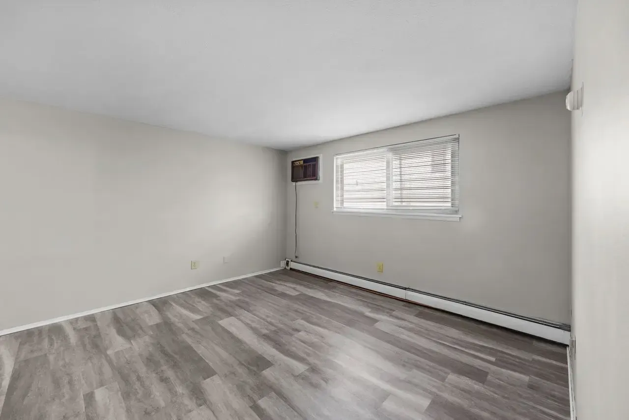An empty room with gray wood-look flooring, light gray walls, a window with closed blinds, and a wall-mounted heater.
