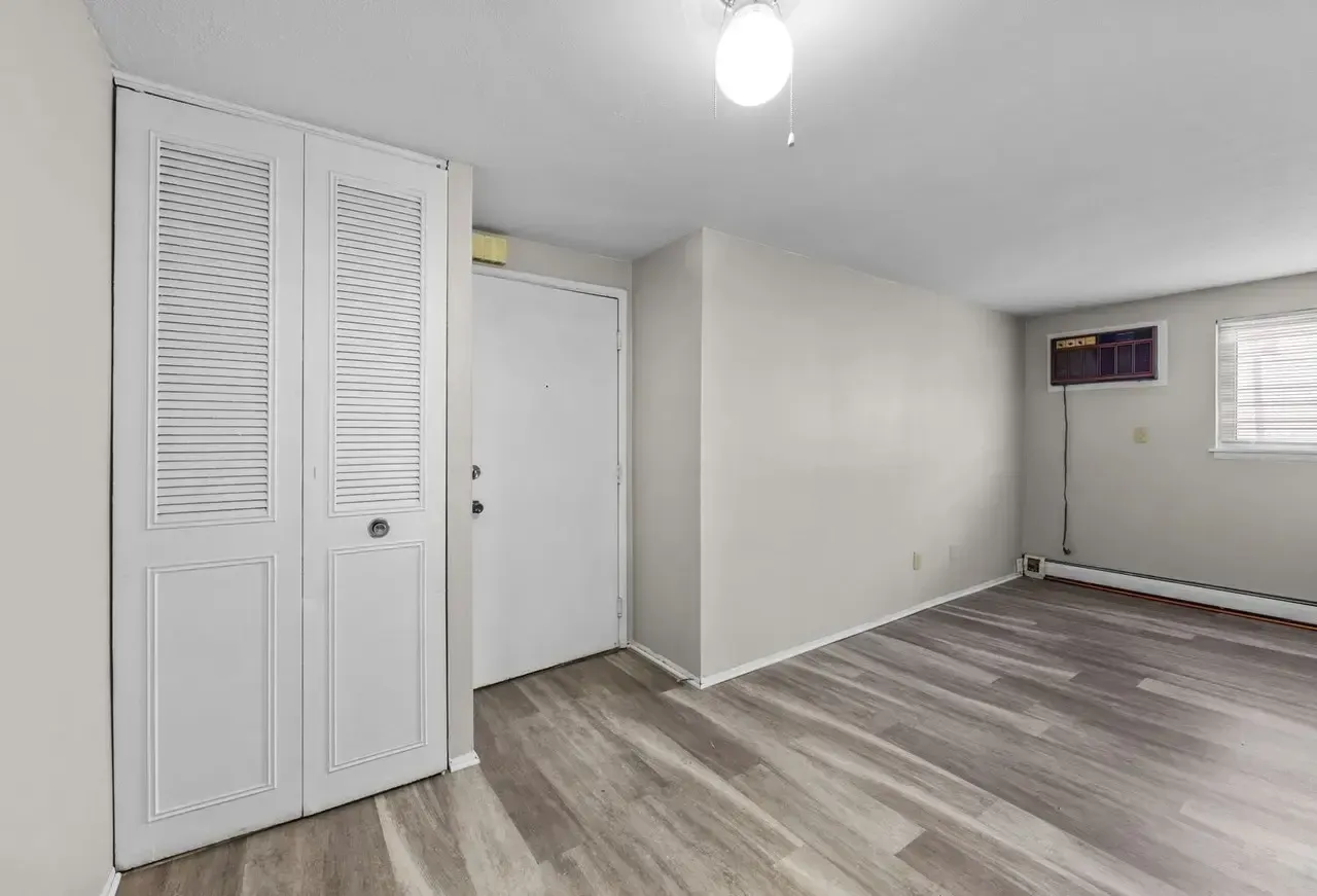 An empty room with light gray walls, wood-look vinyl flooring, white louvered closet doors, a white door, and an AC unit.