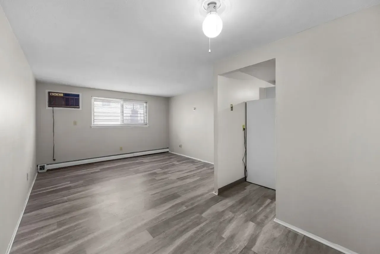 An empty living room with light gray walls, wood-grain flooring, a corner window, and an open doorway to a kitchen.
