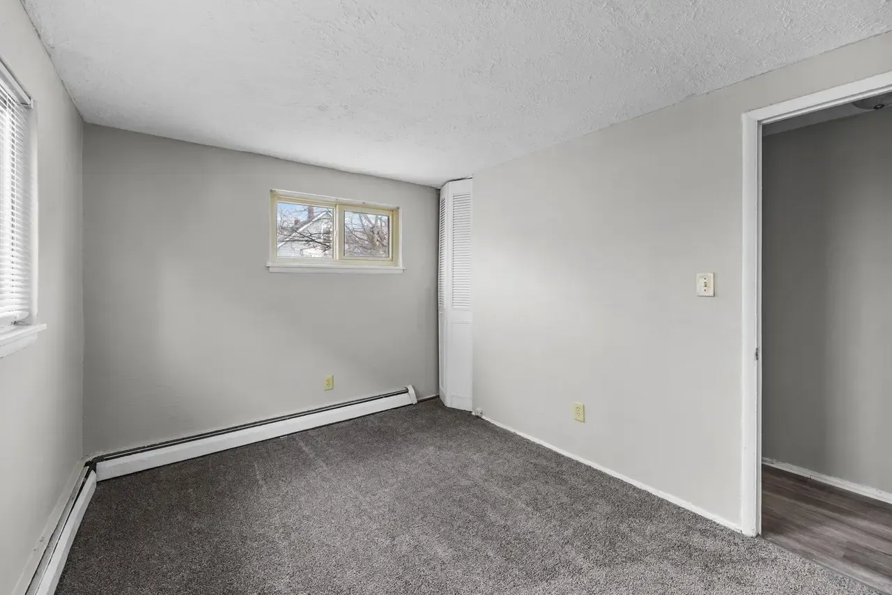 An empty room with gray carpet, light gray walls, a small rectangular window, and a doorway leading to a hallway.