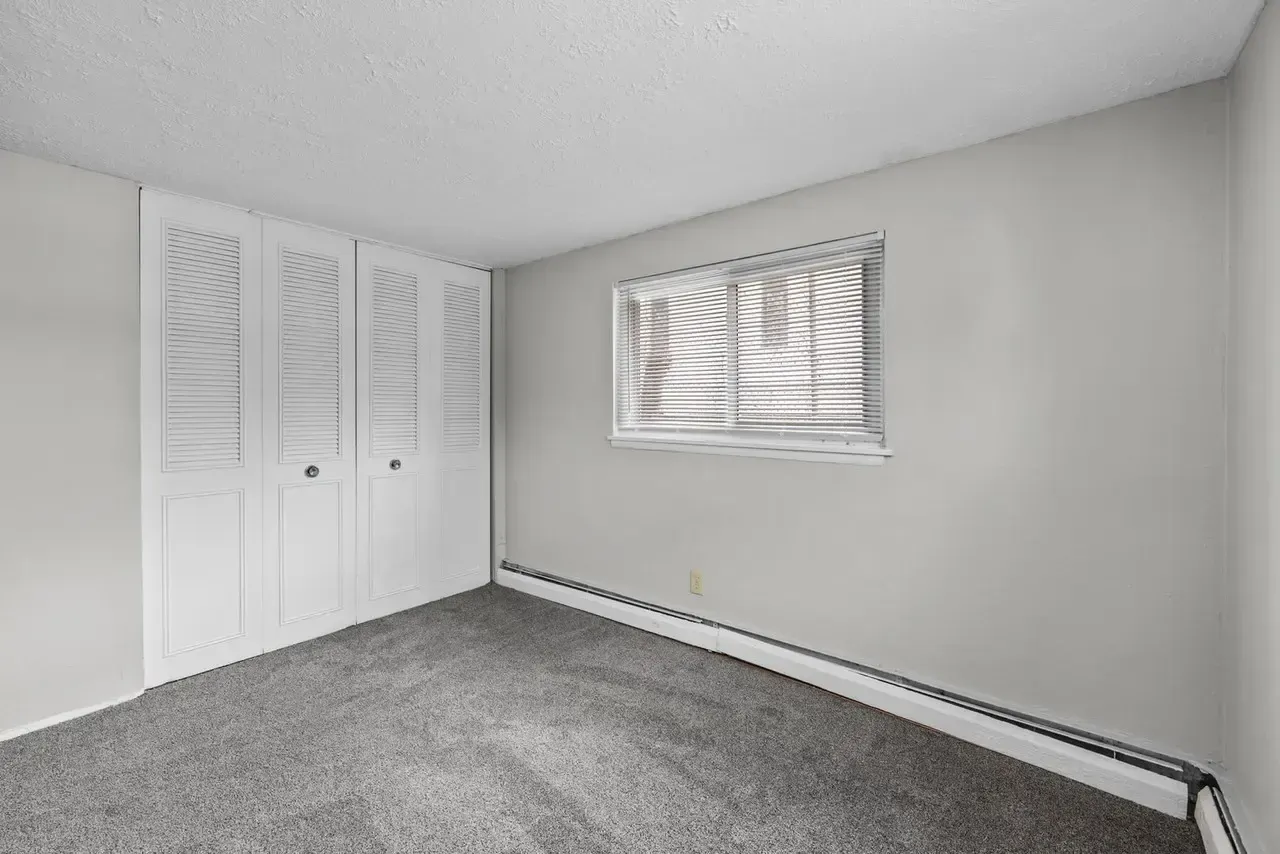 An empty bedroom with light gray walls, gray carpet, a white louvered closet, and a single window with white blinds.