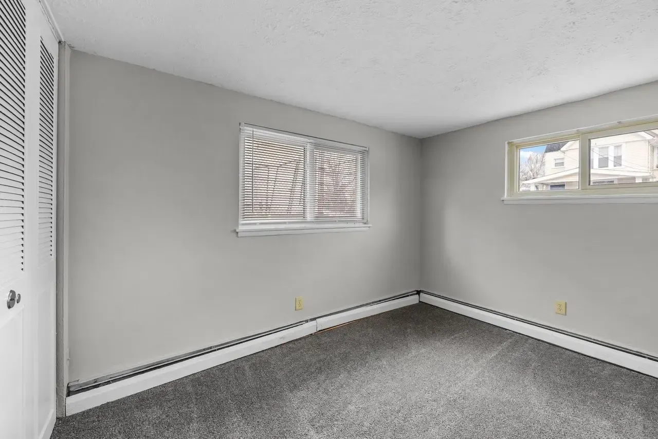 An empty room with light gray walls, gray carpet, baseboard heating, a louvered closet door, and two windows.