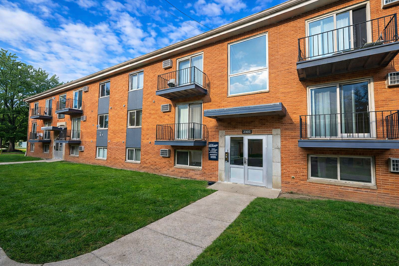 A three-story brick apartment building with black balconies, grey window trim, and a concrete walkway on a grassy lawn.