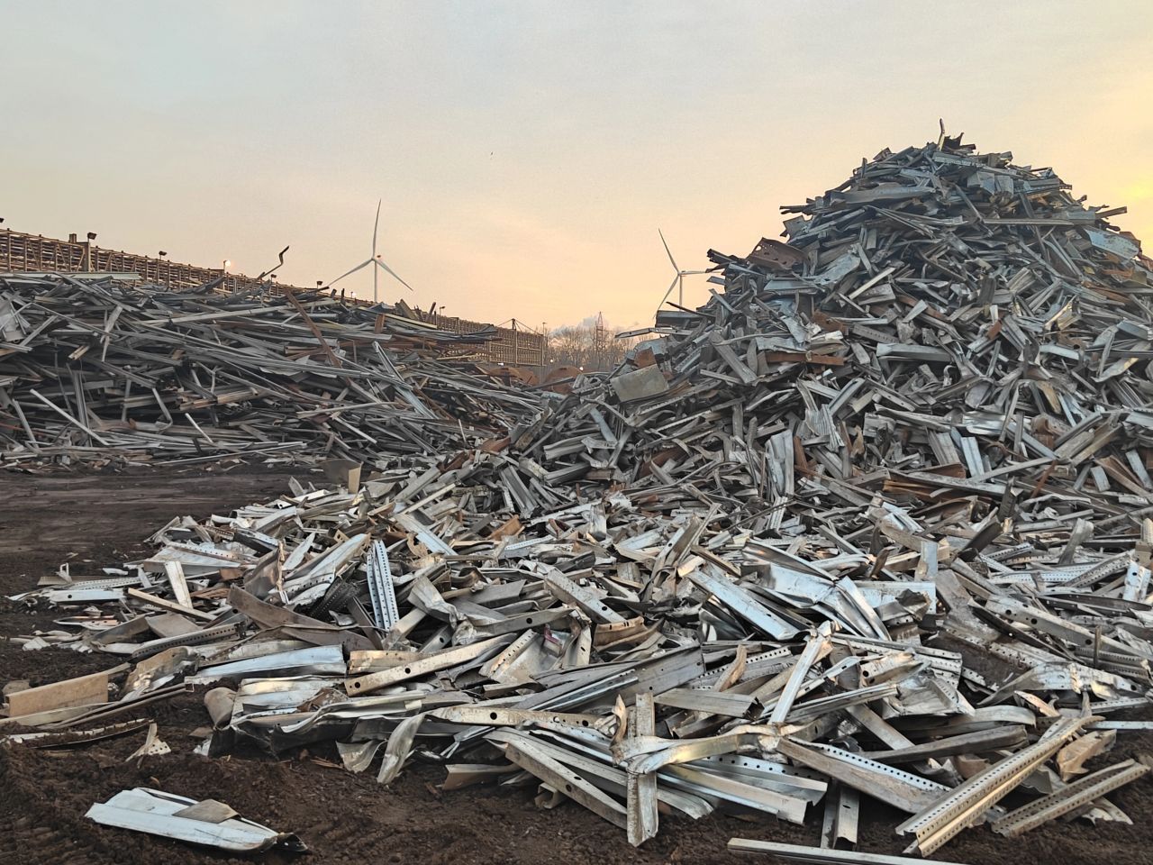 Hopen afvalhout en puin op een kaal veld bij zonsondergang, met windturbines in de verte.