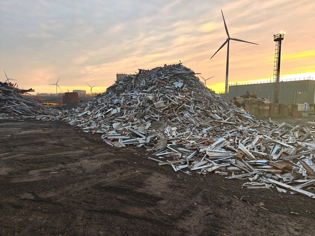 Stapels schroot op een recyclingterrein bij zonsondergang, met een windmolen op de achtergrond.