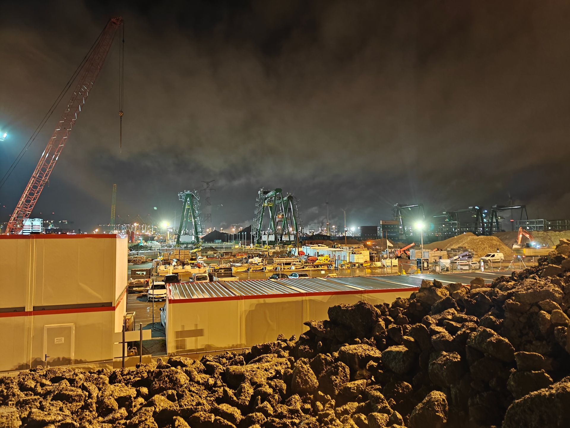 Een bouwplaats 's nachts met oranje containers, een rotsachtige voorgrond en stadslichten onder een bewolkte hemel.