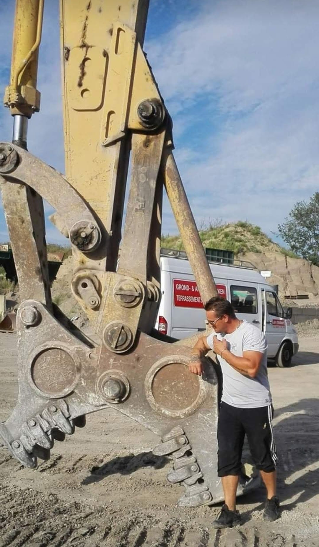 Een man inspecteert de arm van een grote graafmachine op een bouwterrein, met vrachtwagens en grond op de achtergrond.