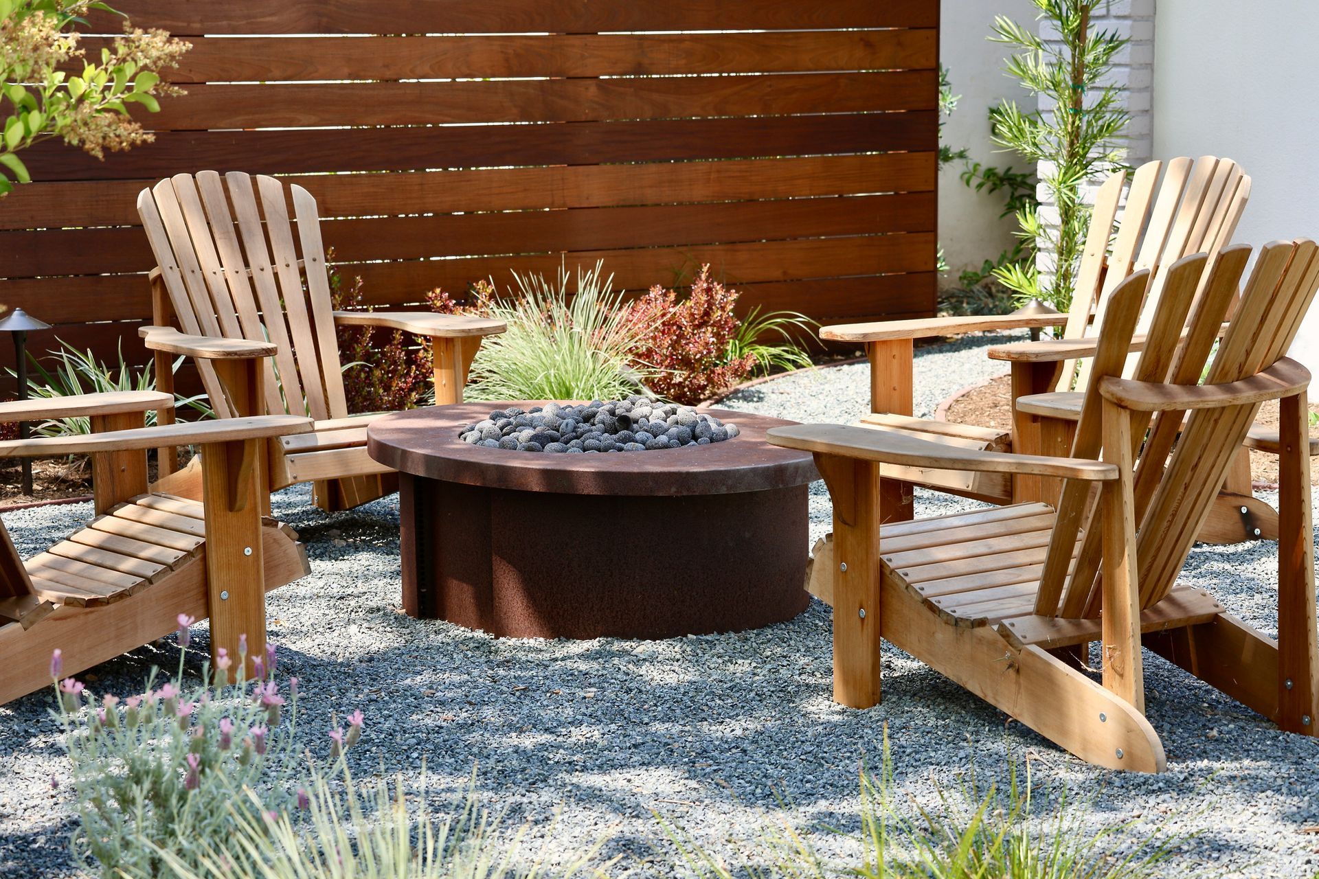 Four wooden Adirondack chairs around a fire pit in a gravel-covered outdoor space.