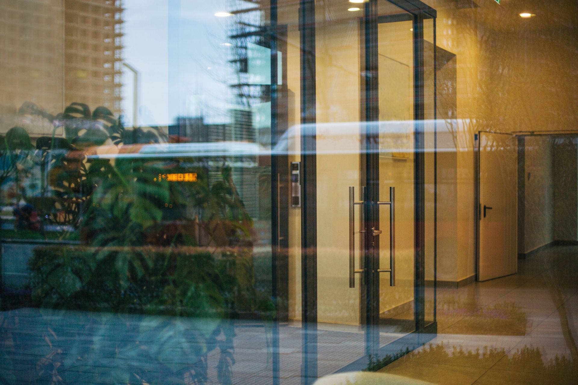 Glass entrance reflecting greenery and a building. Interior shows doors and a hallway with a wet floor. Glass entrance reflecting greenery and a building. Interior shows doors and a hallway with a wet floor.