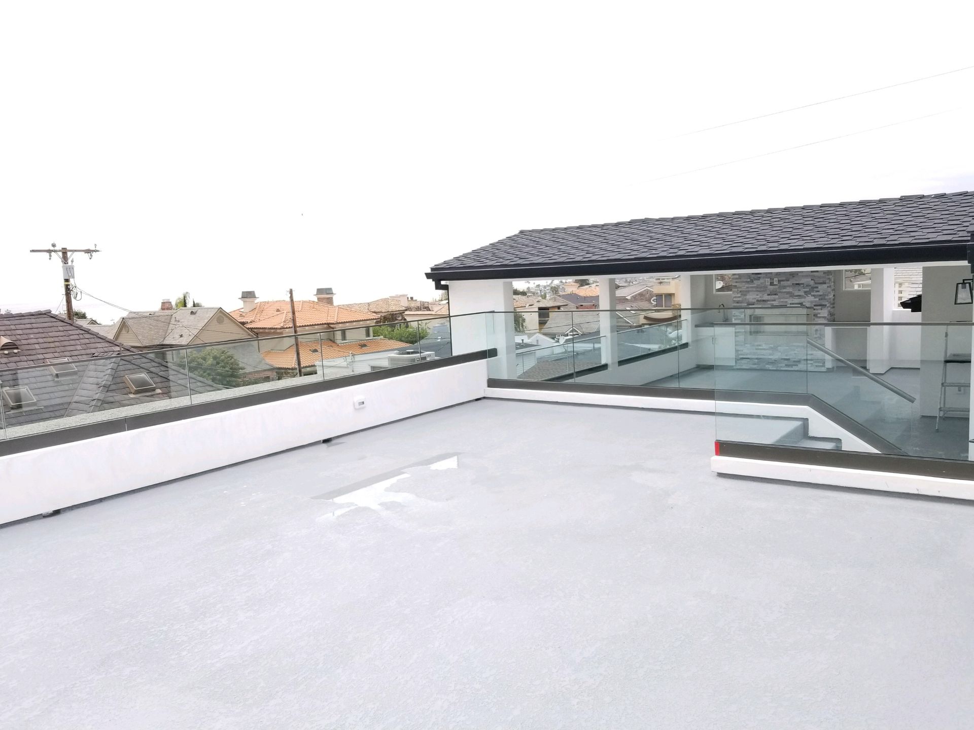 Rooftop with a covered area, glass railings, and a view of houses in the distance under a cloudy sky.