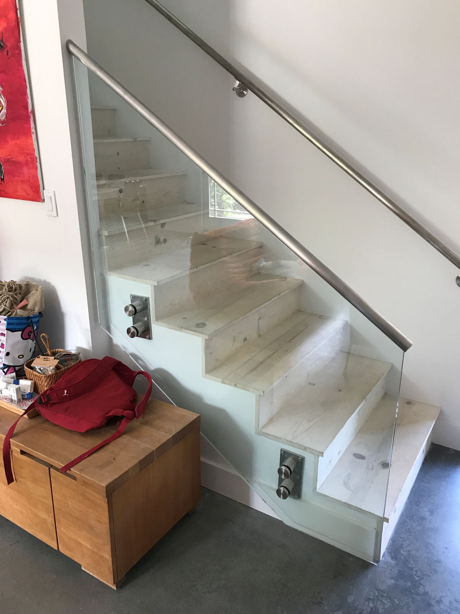 Staircase with glass panel and stainless steel handrail; a wooden trunk and red bag sit at base.