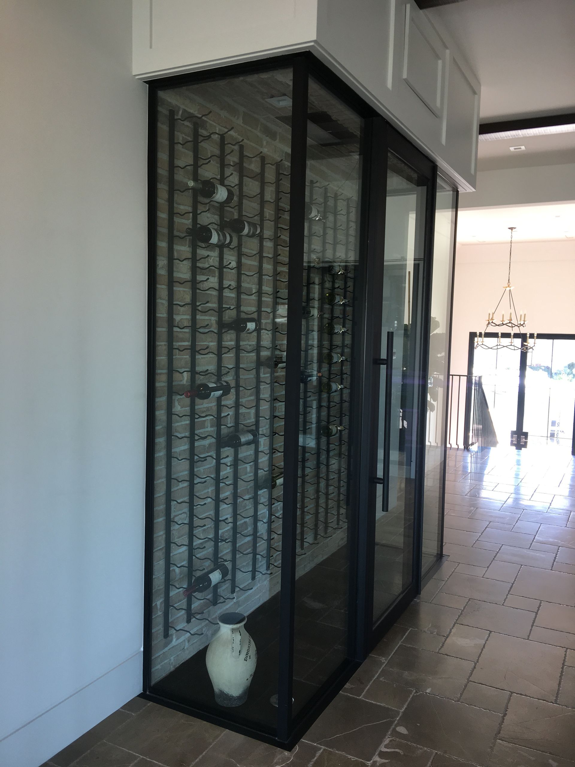 Glass-enclosed wine storage unit with black frame. Bottles are displayed on a brick-patterned back wall, with a vase at the bottom.