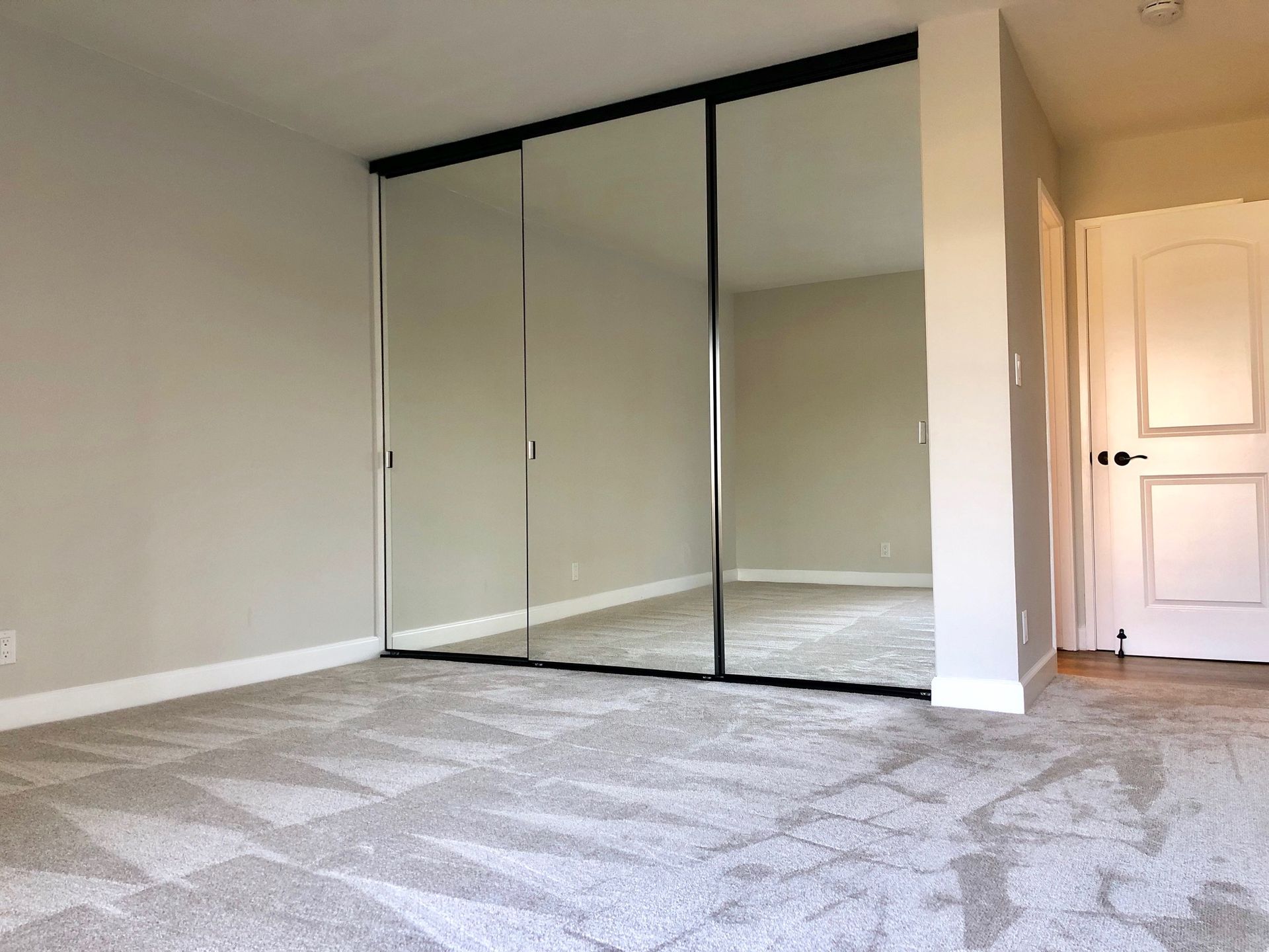 Bedroom with mirrored closet doors and a closed white door on the right. Beige carpet and walls.