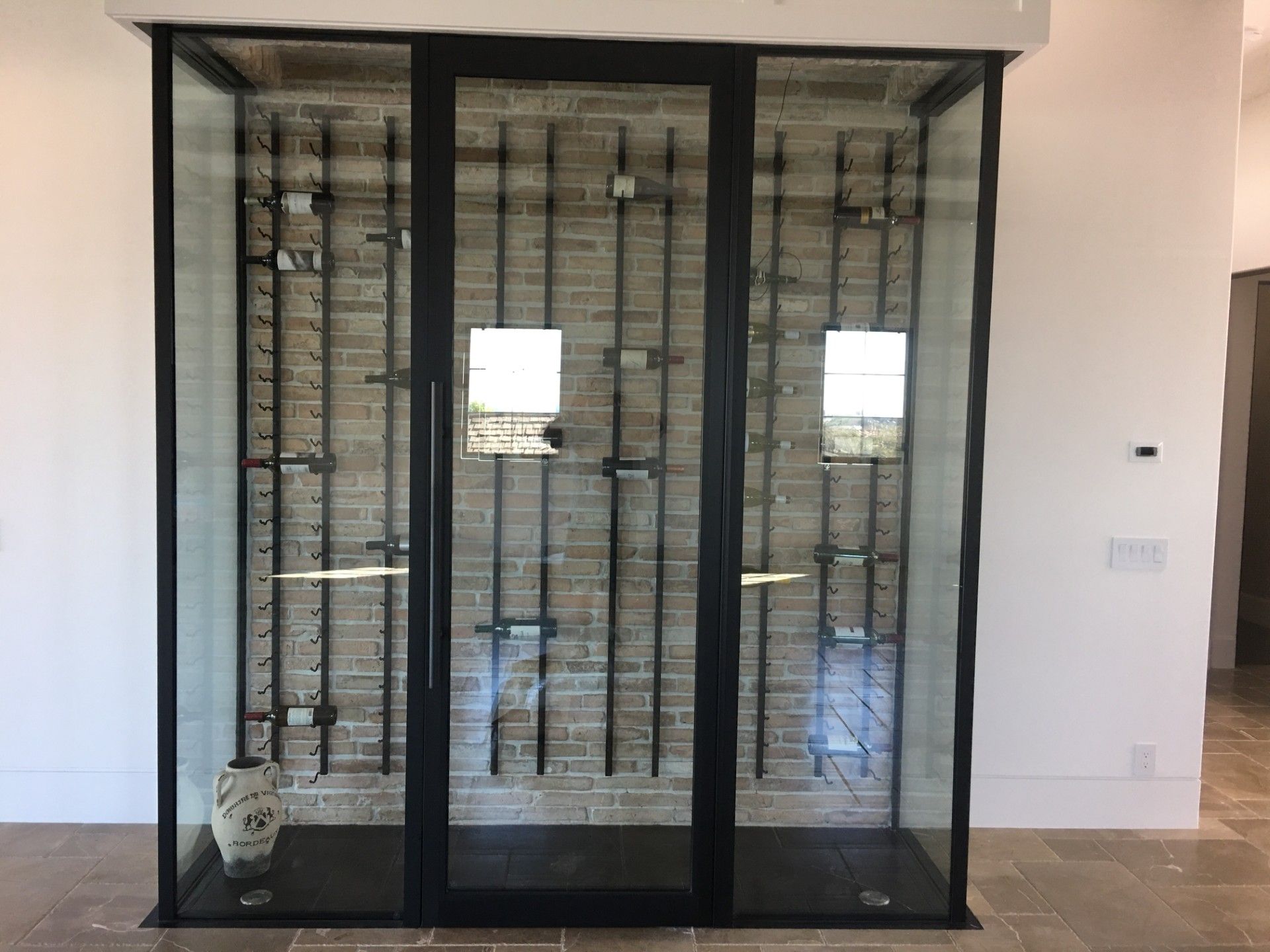 Wine cellar with black metal frame and glass doors; brick backing; bottles on racks.