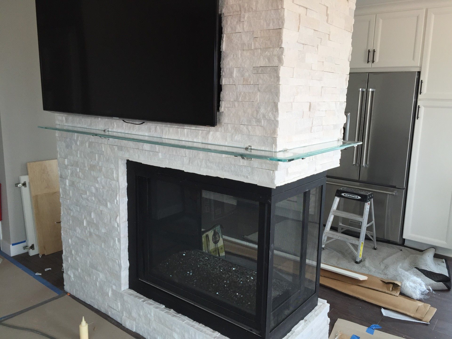 A corner fireplace with white stone and a black-framed glass door. A large TV is mounted above it, and a glass shelf is installed below.