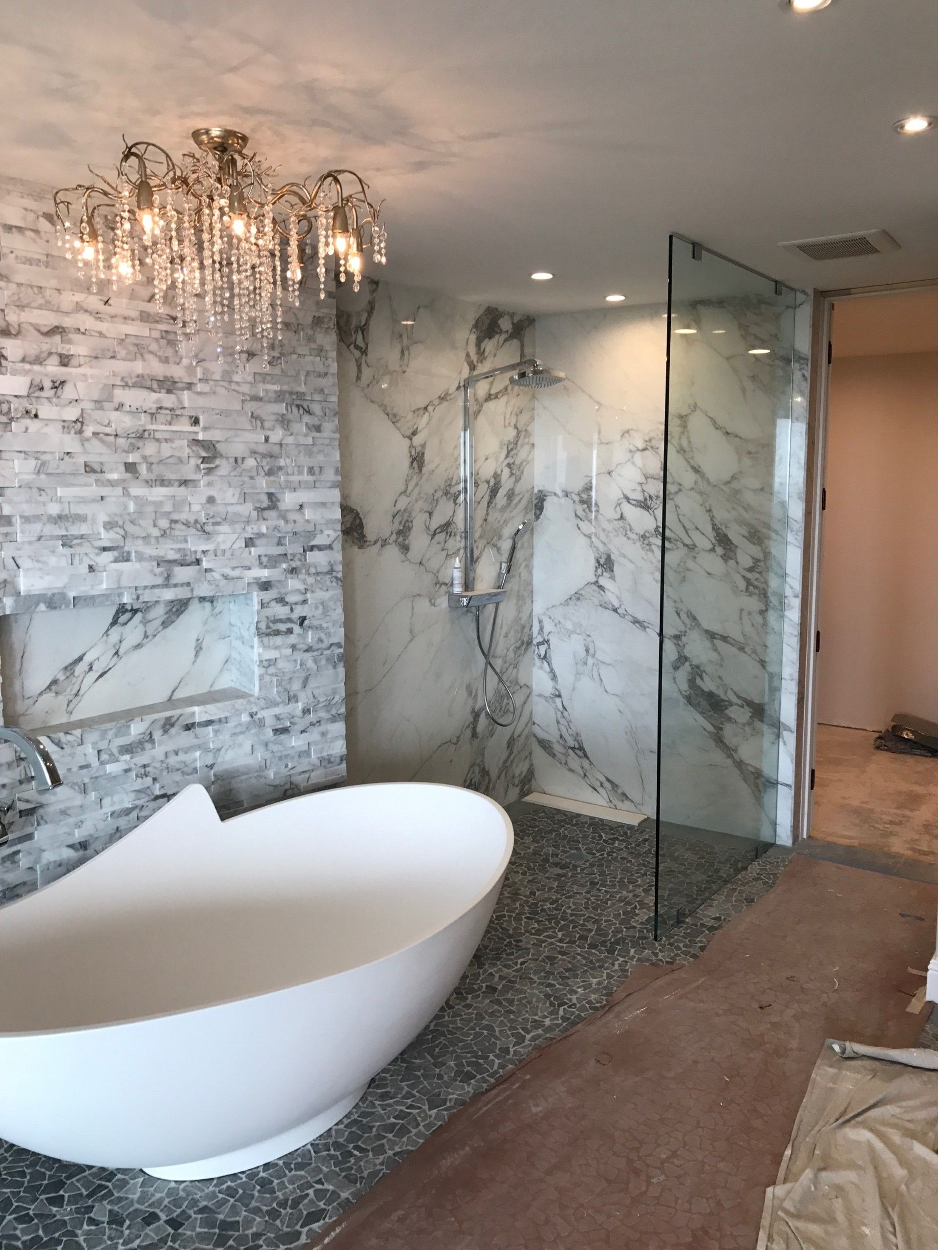 Modern bathroom with white tub, stone shower, chandelier, and marble walls.
