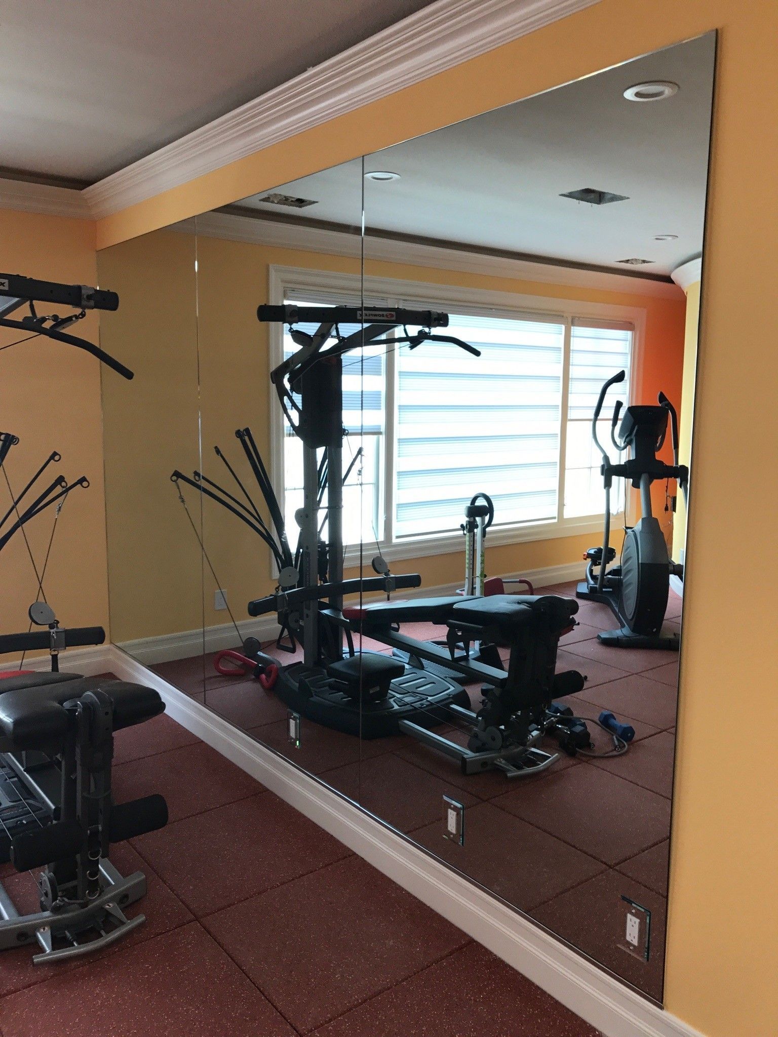 Gym with equipment, reflected in a large wall mirror. Red floor, yellow walls, and a window are visible.