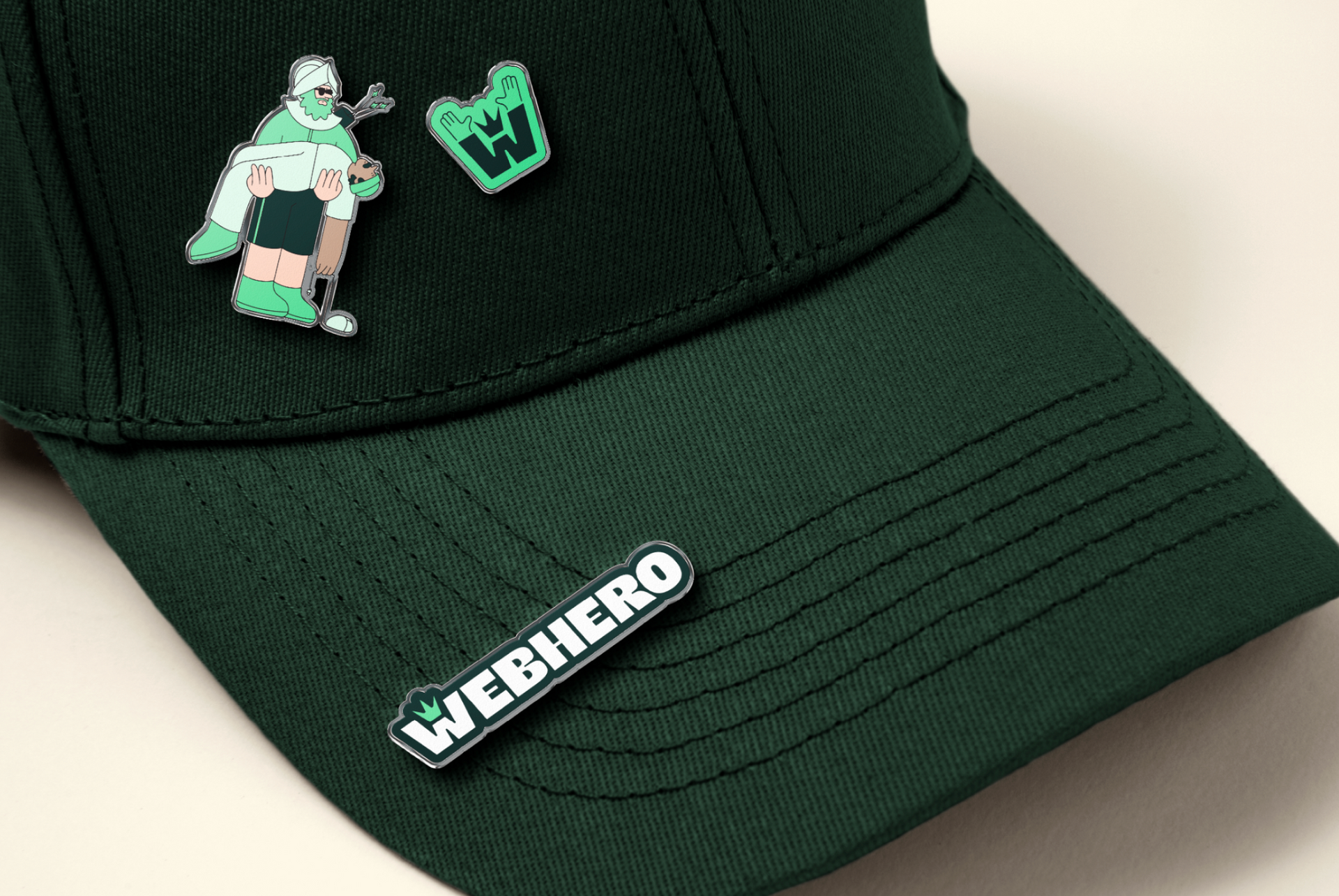 Dark green baseball cap with three pins: pixel art figure, "W" logo, and "WEBHERO" text.