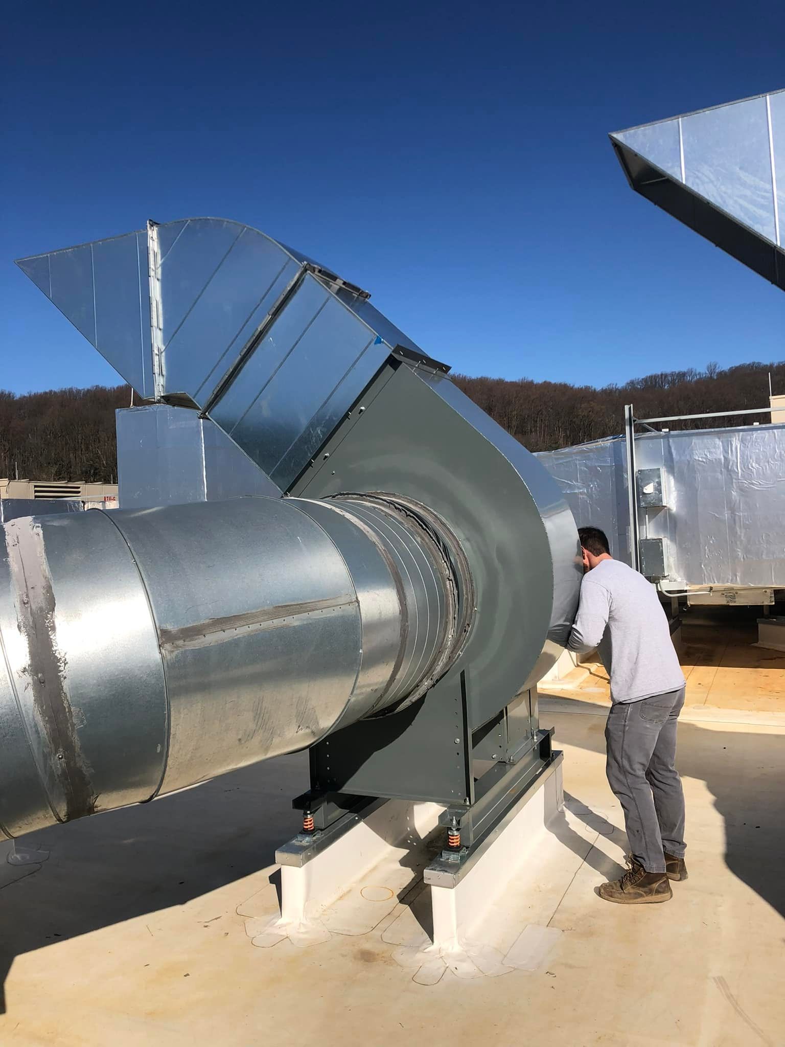 A man is standing next to a large metal fan on a roof.