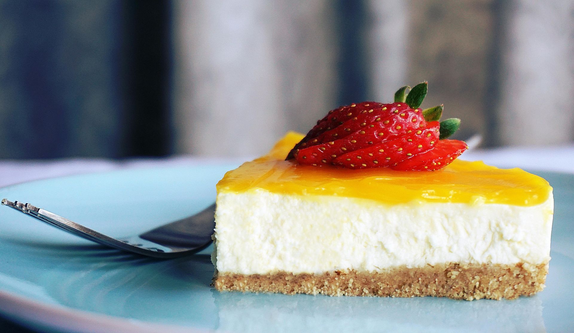 Cheesecake slice on blue plate, topped with yellow glaze and strawberry.