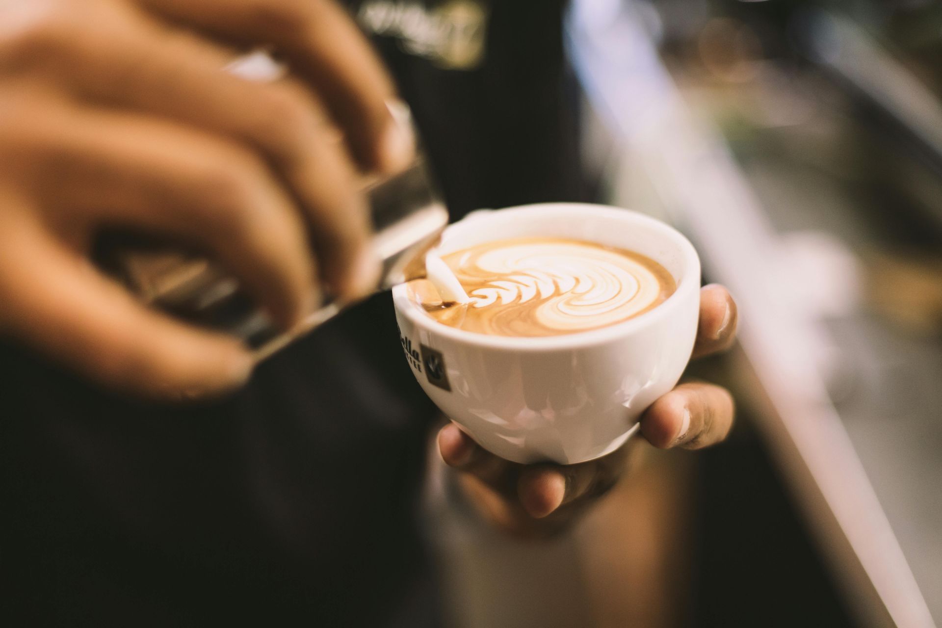 Barista pouring milk, creating latte art in a small white cup. Close-up, warm tones.