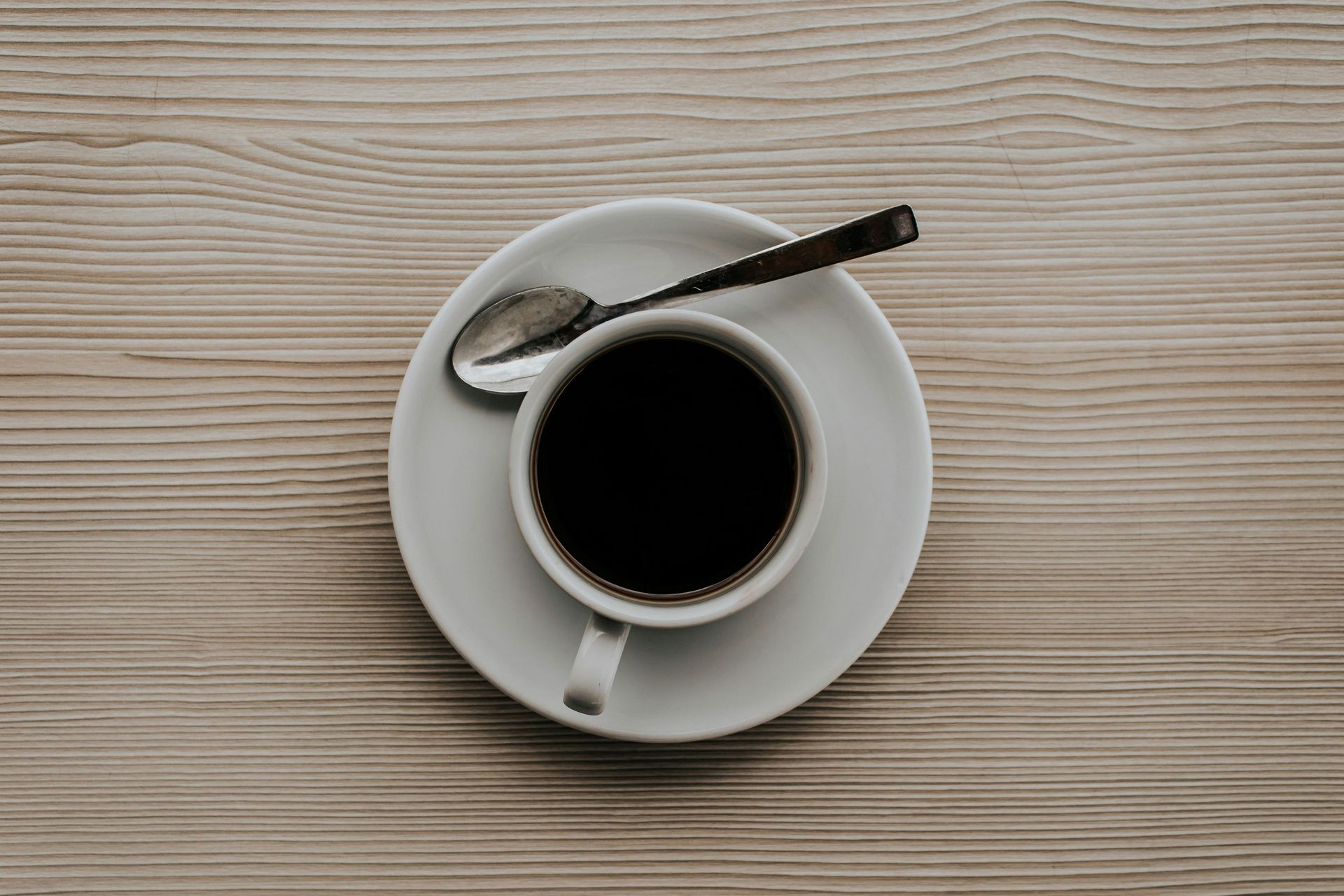 Cup of dark coffee on a white saucer, spoon resting on the saucer, on a wooden surface.