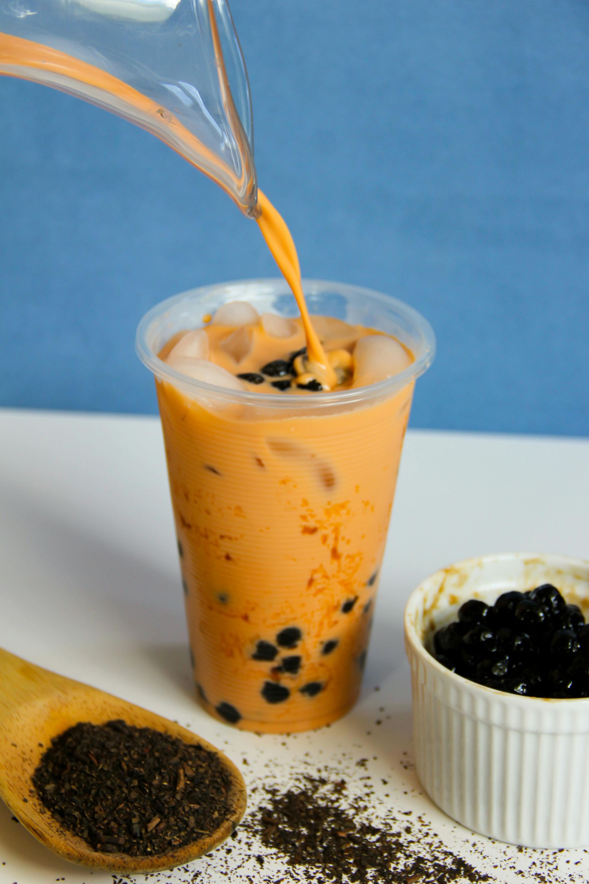 Orange Thai tea being poured into a cup with boba and ice; tea powder and boba are nearby.