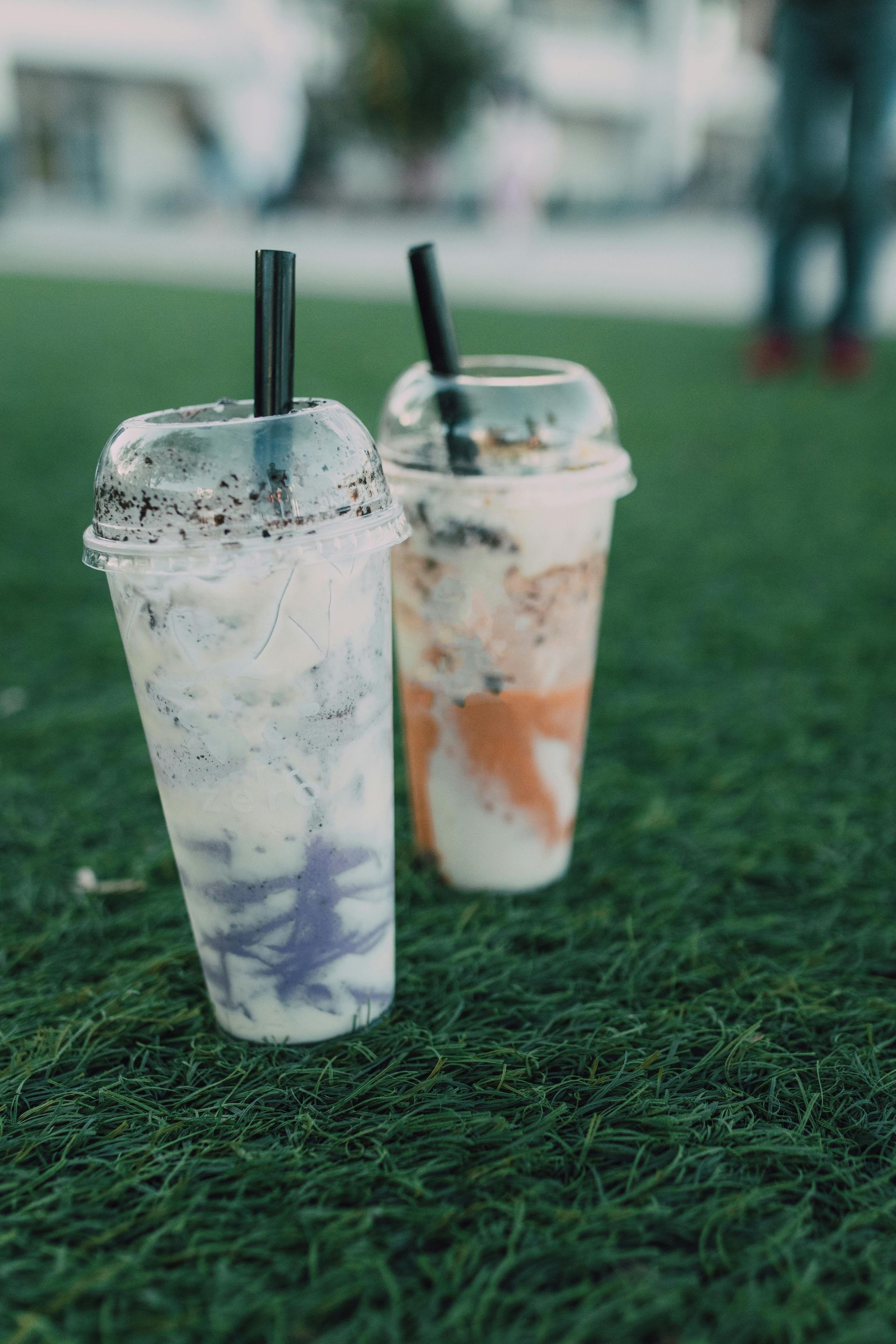 Two colorful drinks in clear plastic cups with black straws on green grass.