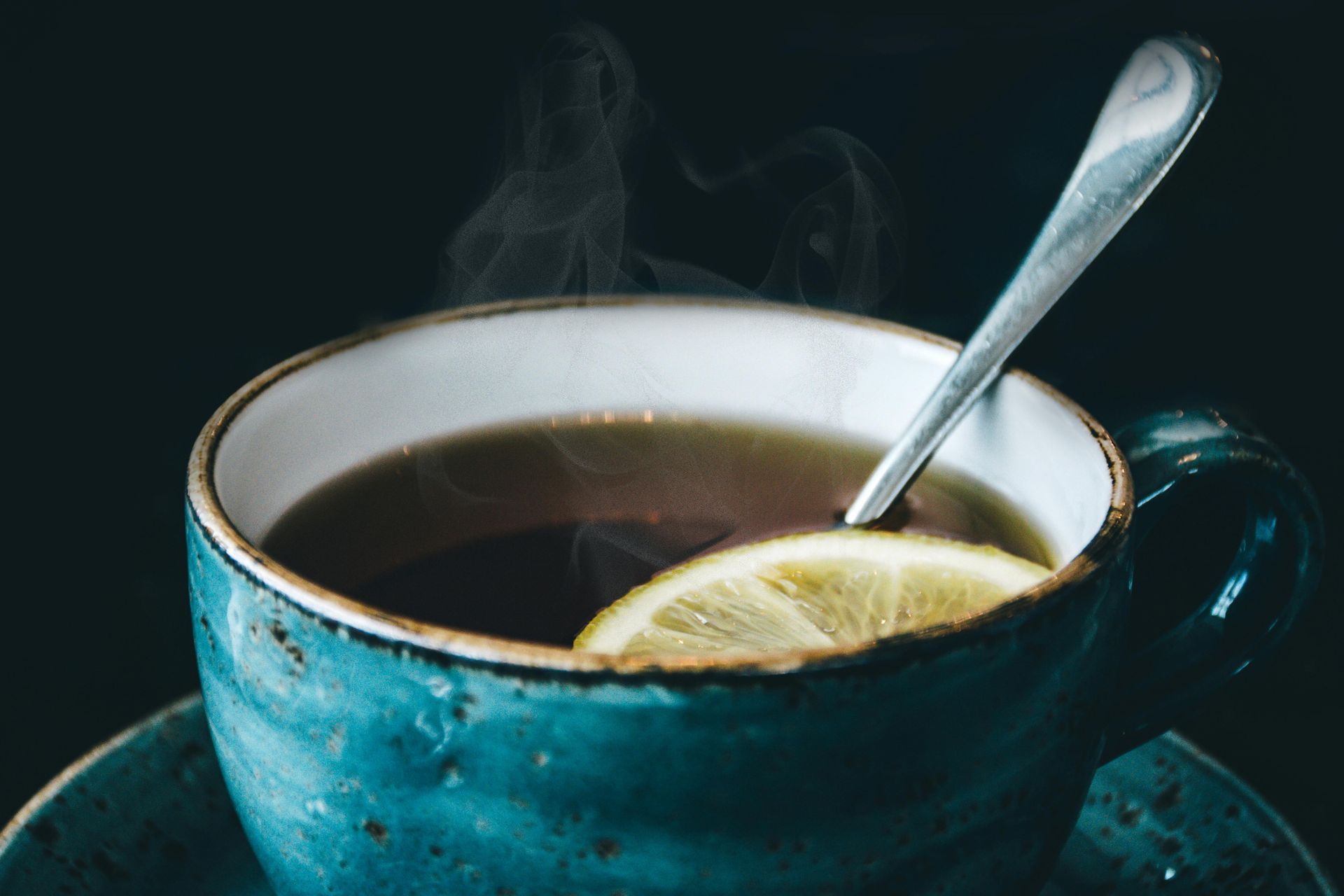 Blue mug of tea with lemon slice, steam rising, spoon resting inside on a dark background.